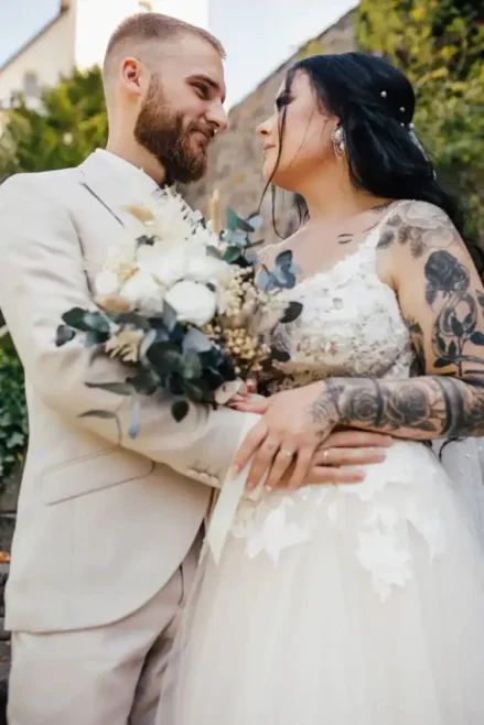 Braut und Bräutigam stehen während ihrer Hochzeit in Tharandt eng beieinander. Die Braut trägt ein Spitzenbrautkleid mit Tattoos und hält einen weißen Blumenstrauß in der Hand. Der Bräutigam trägt einen hellen Anzug. Sie blicken sich in die Augen, umgeben von üppigem Grün im Hintergrund.