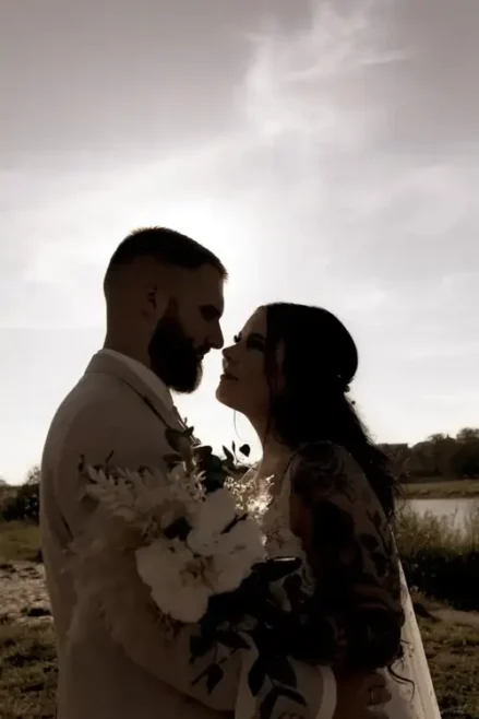 Ein Foto fängt die Romantik einer Hochzeit in Tharandt ein. Das Paar zeichnet sich als Silhouette vor der Sonne ab. Der Mann, bärtig und im Anzug, und die Frau, mit langen Haaren und Kleid, halten einen Blumenstrauß in den Händen, der ihre zeitlose Eleganz unterstreicht.