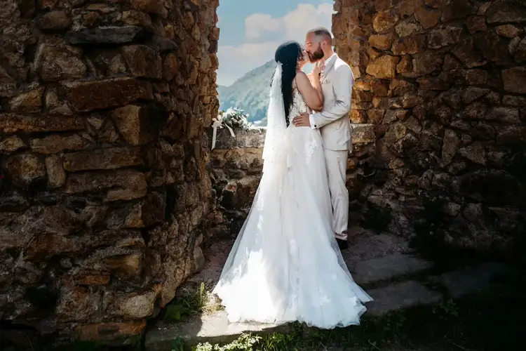 Braut und Bräutigam küssen sich zärtlich inmitten alter Steinmauern, wunderschön eingefangen von Hochzeit in Tharandt-Melanie Kunert Fotografie. Sie trägt ein fließendes weißes Kleid, er einen cremefarbenen Anzug vor der Kulisse malerischer Berge und eines leicht bewölkten Himmels.