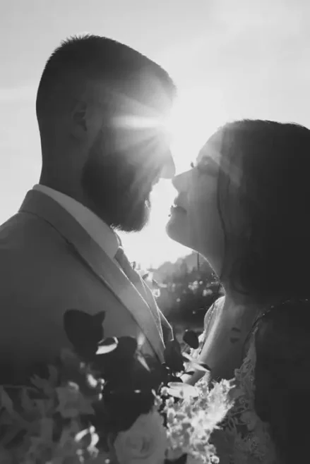 Ein Schwarz-Weiß-Foto fängt die Romantik einer Hochzeit in Tharandt ein. Das Paar zeichnet sich als Silhouette vor der Sonne ab. Der Mann, bärtig und im Anzug, und die Frau, mit langen Haaren und Kleid, halten einen Blumenstrauß in den Händen, der ihre zeitlose Eleganz unterstreicht.