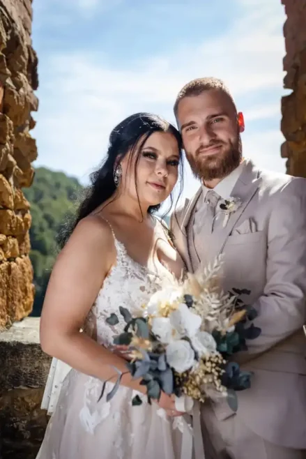 Braut und Bräutigam posieren eng vor einer Steinmauer, wunderschön eingefangen von Melanie Kunert Fotografie bei ihrer Hochzeit in Tharandt. Die Braut hält einen Strauß weißer Blumen und Grünpflanzen in der Hand, während der Bräutigam einen beigen Anzug mit Krawatte trägt, vor der Kulisse von Bäumen und blauem Himmel.