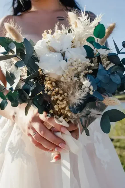 Die Braut steht anmutig da und hält einen Strauß weißer Blumen, Eukalyptusblätter und getrocknetes Laub in den Händen. Sie trägt ein weißes Kleid mit Spitzendetails, das ihre elegante Maniküre und Ringe vor dem Hintergrund eines strahlend blauen Himmels perfekt ergänzt – eine Bilderbuchhochzeit in Tharandt.