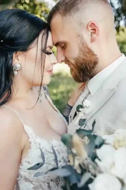 Braut und Bräutigam stehen eng beieinander, die Augen geschlossen, die Stirn berührt sich. Die Braut trägt ein Spitzenkleid, der Bräutigam einen hellen Anzug. Sie hält einen Strauß weißer Blumen und Grünpflanzen in der Hand. Die Szenerie ist im Freien in Tharandt, wunderschön eingefangen von Melanie Kunert Fotografie mit verschwommenen Bäumen im Hintergrund.