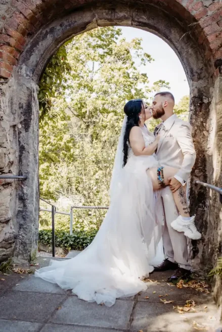 Braut und Bräutigam küssen sich unter einem steinernen Torbogen, wunderschön eingefangen von Melanie Kunert Fotografie. Die Braut in ihrem weißen Kleid und Schleier hebt verspielt ein Bein, während der Bräutigam in einem hellen Anzug daneben steht. Grünes Laub aus Tharandt bildet eine üppige Kulisse für ihren Hochzeitsmoment.
