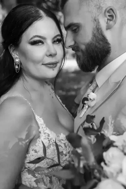 Braut und Bräutigam umarmen sich im Freien bei ihrer Hochzeit in Tharandt, aufgenommen von Melanie Kunert Fotografie. Die Braut trägt ein weißes Spitzenkleid mit einem Strauß weißer und blauer Blumen, während der Bräutigam in einem hellen Anzug sanft ihre Taille inmitten üppigen Grüns hält, das in sanftes, natürliches Licht getaucht ist.