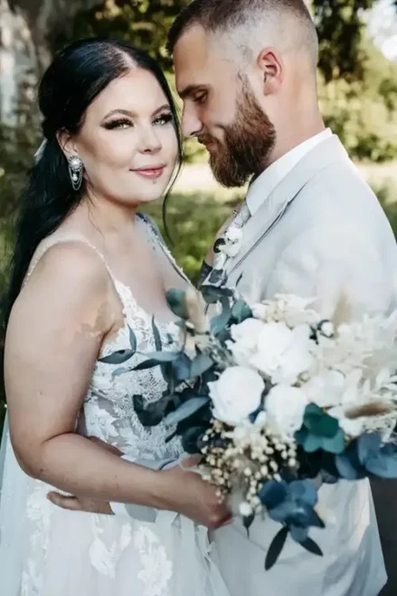 Braut und Bräutigam umarmen sich im Freien bei ihrer Hochzeit in Tharandt, aufgenommen von Melanie Kunert Fotografie. Die Braut trägt ein weißes Spitzenkleid mit einem Strauß weißer und blauer Blumen, während der Bräutigam in einem hellen Anzug sanft ihre Taille inmitten üppigen Grüns hält, das in sanftes, natürliches Licht getaucht ist.