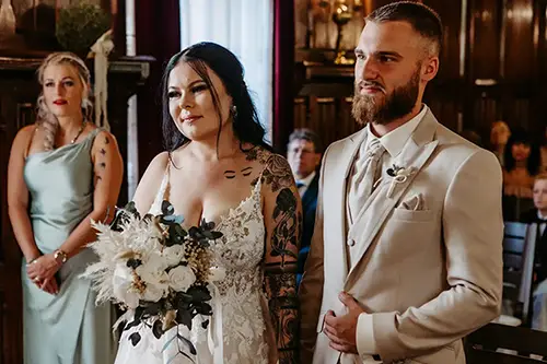 Ein Paar steht in einem warm beleuchteten Raum zusammen, in Hochzeitskleidung, wunderschön eingefangen von Hochzeit in Tharandt-Melanie Kunert Fotografie. Die Braut hält in ihrem weißen Spitzenkleid einen Blumenstrauß, während der Bräutigam einen hellen Anzug trägt. Eine Braut in einem grünen Kleid beobachtet die Zeremonie.