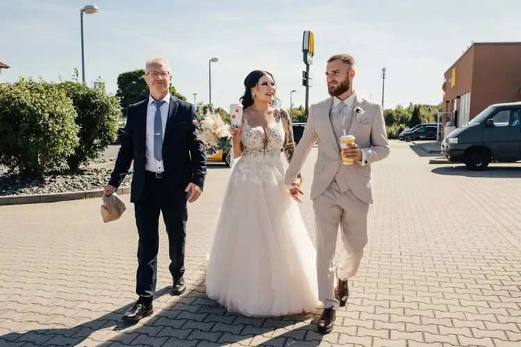 Eine Braut im weißen Kleid und ein Bräutigam im beigen Anzug feiern ihre Hochzeit in Tharandt Hand in Hand mit einem Mann im Anzug und einer Tasche in der Hand. Der Bräutigam genießt einen Drink, während sie über den Parkplatz schlendern, umgeben von Gebäuden und Grünflächen.