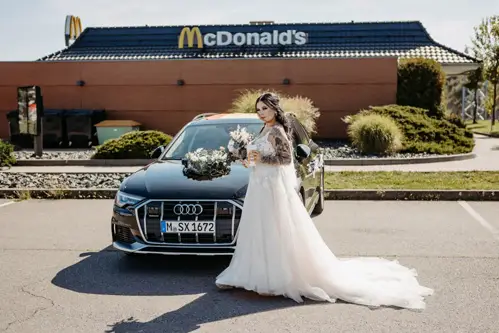 Eine Braut im weißen Kleid steht neben einem schwarzen, mit Blumen geschmückten Audi, der vor einem McDonald's-Restaurant parkt. Unter dem klaren Himmel hält sie einen Blumenstrauß in der Hand. Sie feiert damit ihre Hochzeit in Tharandt. Im Hintergrund ist das McDonald's-Gebäude zu sehen.