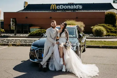 Braut und Bräutigam sitzen lächelnd auf der Motorhaube eines schwarzen Autos. Die Braut, in weißem Kleid und Turnschuhen, und der Bräutigam, in hellem Anzug, posieren an einem sonnigen Tag vor einem McDonald's-Restaurant in Tharandt für ihre Hochzeit.