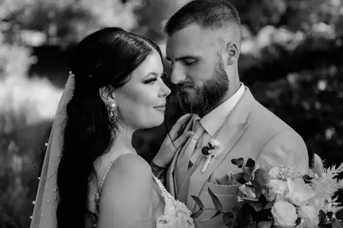 Ein Schwarz-Weiß-Foto zeigt Braut und Bräutigam in intimer Pose während ihrer Hochzeit in Tharandt. Die Braut hält einen Blumenstrauß in der Hand und blickt den Bräutigam an, der leicht lächelt, während die beiden in festlicher Kleidung vor üppigem Laub stehen.