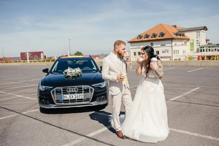 Auf einem sonnigen Parkplatz teilen sich Braut und Bräutigam Fast Food vor einem schwarzen, mit weißen Blumen geschmückten Audi. Fröhlich lächelnd feiern die Braut in ihrem weißen Kleid und der Bräutigam im hellen Anzug ihre Hochzeit in Tharandt.