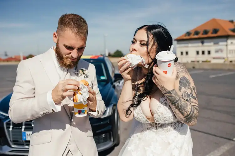 Braut und Bräutigam in Hochzeitskleidung genießen Fast-Food-Burger und halten Getränke vor einem geparkten Auto, im Hintergrund ragen die Gebäude empor. Die tätowierte Braut beißt begeistert in ihren Burger und hält damit einen einzigartigen Moment der Hochzeit in Tharandt fest.