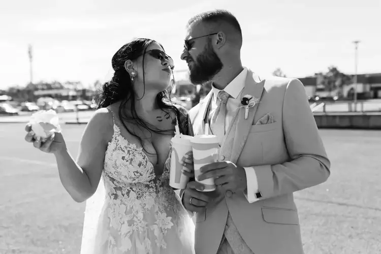 Eine Braut im weißen Spitzenkleid und ein Bräutigam im beigen Anzug lachen und halten draußen auf einem Tharandter Parkplatz Fastfood in der Hand. Fröhlich, mit Sonnenbrille und vom Wind zerzaustem Haar verkörpern sie den Geist ihres Hochzeitsabenteuers.