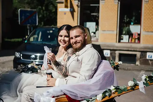 Ein lächelndes Paar in Hochzeitskleidung sitzt während seiner Hochzeit in Tharandt in einem geschmückten Wagen. Die Braut im weißen Kleid hält eine Flasche, während der Bräutigam im hellen Anzug neben ihr strahlt. Sie stehen im Freien, hinter ihnen sind ein Auto und ein Gebäude zu sehen.