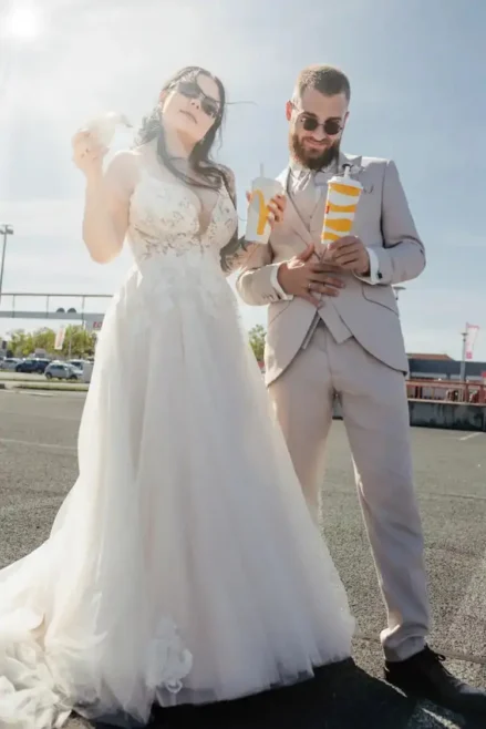 Inmitten der lebhaften Atmosphäre von Hochzeit in Tharandt stehen eine Braut in einem weißen Kleid und einer Sonnenbrille und ein Bräutigam in einem beigen Anzug stilvoll auf dem Bürgersteig und genießen Getränke und Snacks unter einem klaren blauen Himmel, über dem die Sonne hell scheint.