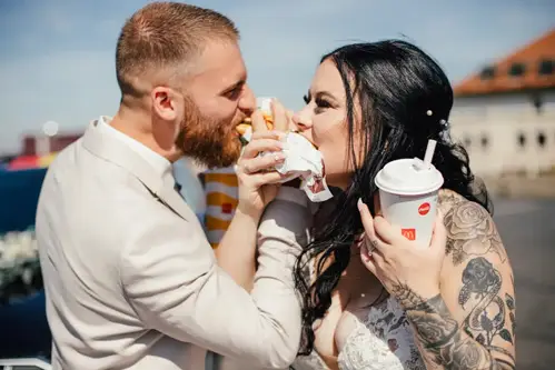 Braut und Bräutigam in Hochzeitskleidung genießen während ihrer Hochzeit einen Burger und Limonade. Der Bräutigam trägt einen hellen Anzug, während die tätowierte Braut in ihrem weißen Kleid strahlt. Ihre Lächeln strahlen und strahlen pure Freude vor der bezaubernden Kulisse aus.