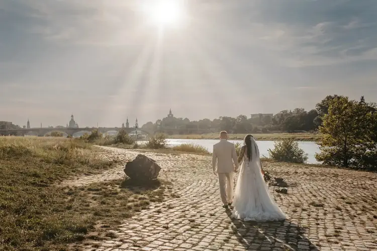 Ein Paar in Hochzeitskleidung hält Händchen auf einem Kopfsteinpflasterweg an einem Fluss. Im Hintergrund ist die Silhouette von Dresden zu sehen. Der Himmel ist klar und sonnig und wirft ein sanftes Licht auf die Hochzeitsszene.