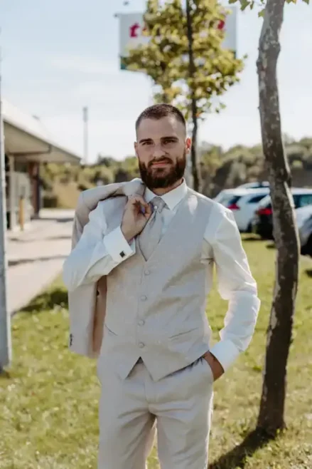 Ein Mann mit Bart steht an einem sonnigen Tag im Freien. Er trägt einen beigen Anzug und eine Weste. Eine Hand in der Tasche, die andere lässig die Jacke über der Schulter, verströmt er Charme, als wäre er bereit für eine Hochzeit in Tharandt, vor der Kulisse parkender Autos und Bäume.