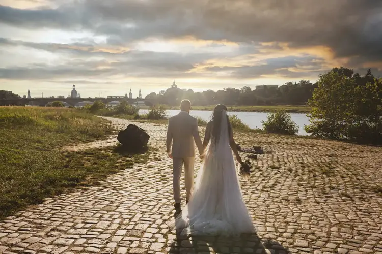 Ein Paar in Hochzeitskleidung hält Händchen auf einem Kopfsteinpflasterweg an einem Fluss. Im Hintergrund ist die Silhouette von Dresden zu sehen. Der Himmel ist klar und sonnig und wirft ein sanftes Licht auf die Hochzeitsszene.