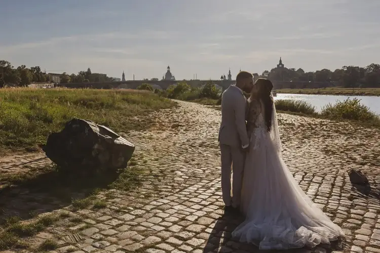 Ein Paar in Hochzeitskleidung hält Händchen auf einem Kopfsteinpflasterweg an einem Fluss. Im Hintergrund ist die Silhouette von Dresden zu sehen. Der Himmel ist klar und sonnig und wirft ein sanftes Licht auf die Hochzeitsszene.