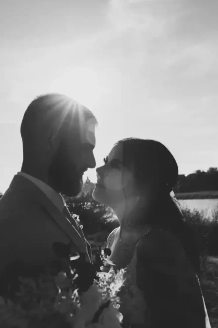 Ein Schwarz-Weiß-Foto fängt die Romantik einer Hochzeit in Tharandt ein. Das Paar zeichnet sich als Silhouette vor der Sonne ab. Der Mann, bärtig und im Anzug, und die Frau, mit langen Haaren und Kleid, halten einen Blumenstrauß in den Händen, der ihre zeitlose Eleganz unterstreicht.