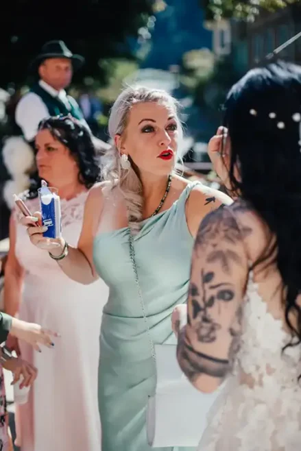 Bei einer Hochzeit in Tharandt schminkt eine Frau in einem hellblauen Kleid die Braut nach und hält ein Getränk in der Hand. Sie steht mit mehreren Personen im Freien, darunter eine tätowierte Person in einem weißen Kleid. Sonnenlicht fällt durch die Bäume und schafft eine helle, lebendige Atmosphäre.