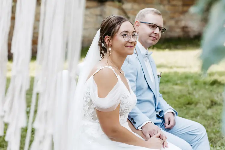 Braut und Bräutigam, gekleidet in ein weißes Spitzenbrautkleid und einen hellblauen Anzug, sitzen Händchen haltend auf Stühlen. Der Anzug des Bräutigams ist mit einem Einstecktuch und einer Ansteckblume geschmückt. Diese ruhige Zeremonie im Freien auf einer Wiese wurde von Melanie Kunert Fotografie, der renommierten Hochzeitsfotografin Dresden, wunderschön eingefangen.