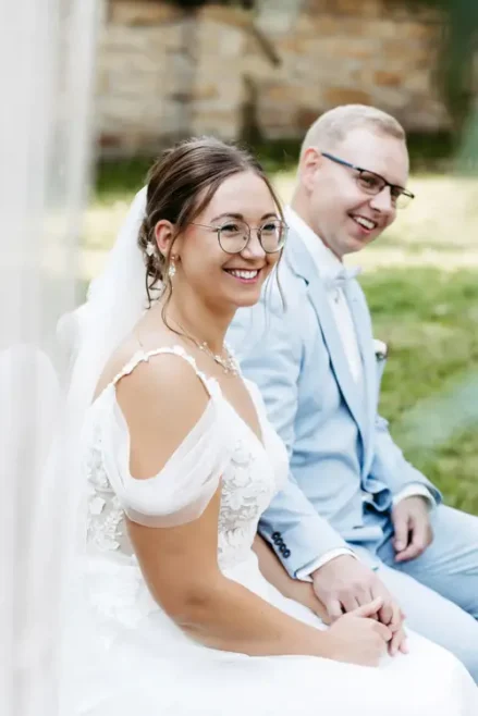 Braut und Bräutigam, gekleidet in ein weißes Spitzenbrautkleid und einen hellblauen Anzug, sitzen Händchen haltend auf Stühlen. Der Anzug des Bräutigams ist mit einem Einstecktuch und einer Ansteckblume geschmückt. Diese ruhige Zeremonie im Freien auf einer Wiese wurde von Melanie Kunert Fotografie, der renommierten Hochzeitsfotografin Dresden, wunderschön eingefangen.