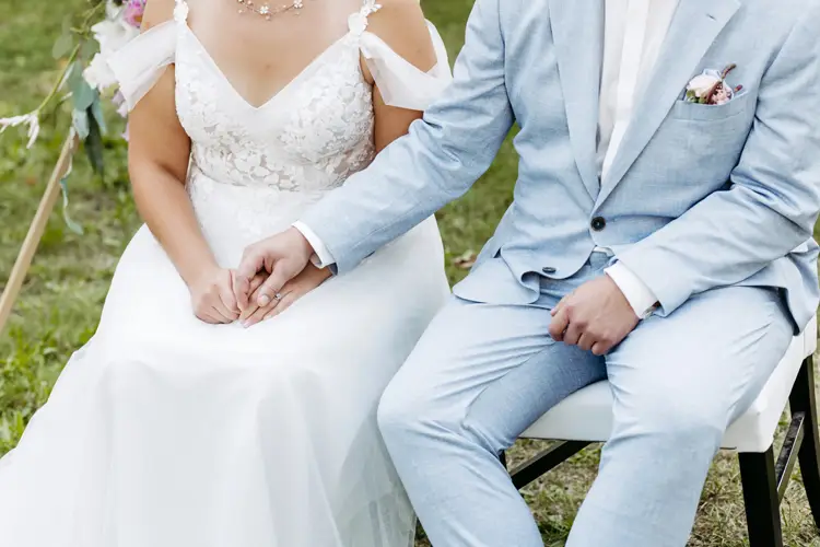 Braut und Bräutigam, gekleidet in ein weißes Spitzenbrautkleid und einen hellblauen Anzug, sitzen Händchen haltend auf Stühlen. Der Anzug des Bräutigams ist mit einem Einstecktuch und einer Ansteckblume geschmückt. Diese ruhige Zeremonie im Freien auf einer Wiese wurde von Melanie Kunert Fotografie, der renommierten Hochzeitsfotografin Dresden, wunderschön eingefangen.