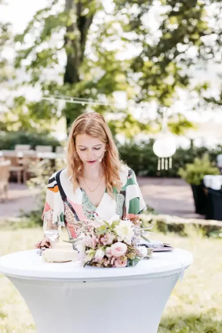 Die Traurednerin mit hellem Haar steht an einem kleinen runden Tisch im Freien. Sie trägt ein farbenfrohes Kleid und konzentriert sich auf die Aktivitäten vor ihr. Auf dem Tisch steht ein Blumenstrauß, im Hintergrund sind Bäume und Tische sanft verschwommen.