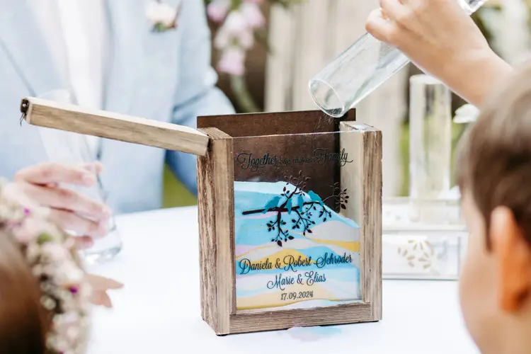 Eine Holzkiste mit der Gravur „Together We Make a Family“ auf dem Deckel enthält eine dekorative Glasplatte mit einem Baummotiv und Daten. In der nahegelegenen Marcolinis Welt nehmen Gäste an einer Sandzeremonie für eine Hochzeit teil, die vom erfahrenen Blick des Hochzeitsfotografen festgehalten wurde.