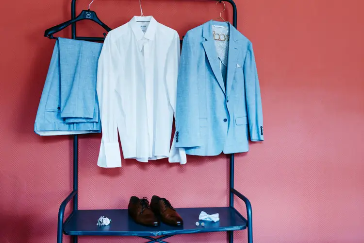 Ein hellblaues Sakko und eine hellblaue Anzughose, ein weißes Hemd, braune Schuhe, eine blaue Fliege und Manschettenknöpfe werden elegant auf einem blauen Kleiderständer vor einer roten Wand präsentiert. Dieses Ensemble eignet sich ideal für Fotos eines Hochzeitsfotografen im Hotel Rothenburger Hof.