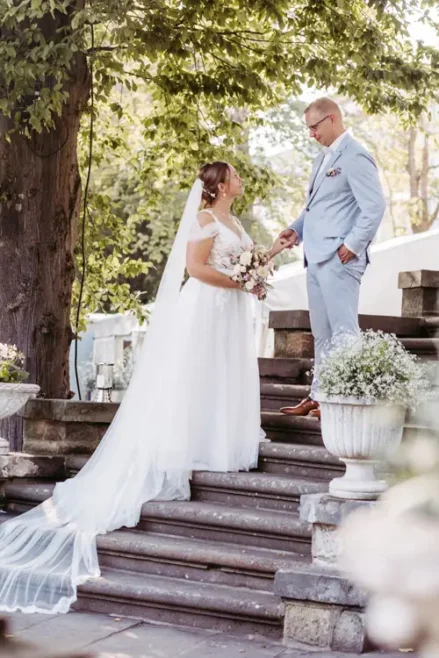 Eine Braut in einem fließenden weißen Kleid steht mit einem Blumenstrauß in der Hand auf einer Steintreppe und tauscht mit dem Bräutigam im hellblauen Anzug das Eheversprechen aus. Umgeben von üppigem Grün und Blumentöpfen wurde diese ruhige Szenerie im Freien vom Hochzeitsfotografen in Marcolinis Welt perfekt eingefangen.