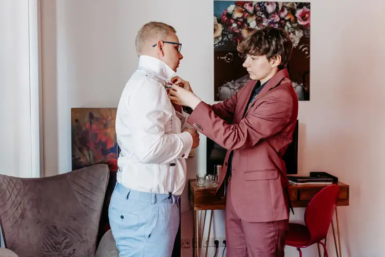 In einer Szene fotografiert der Hochzeitsfotograf zwei Männer in formeller Kleidung. Einer trägt ein frisches weißes Hemd und der andere Herr im Anzug hilft ihm beim Zurechtrücken seines Kragens. Hinter ihnen hängt das Porträt einer Frau mit Blumenkopfschmuck.