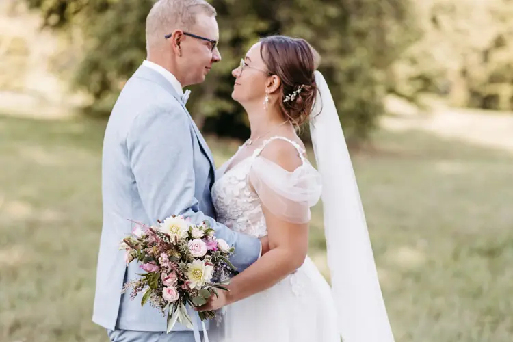 Braut und Bräutigam stehen sich Händchen haltend in einer sonnigen Außenkulisse gegenüber, aufgenommen von einem Hochzeitsfotografen aus Dresden. Die Braut trägt ein weißes Kleid mit langem Schleier, der Bräutigam einen hellgrauen Anzug. Umgeben von Gras und Bäumen entsteht eine friedliche Atmosphäre.