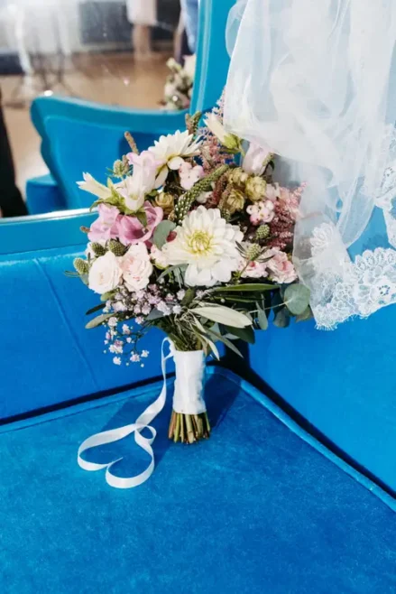 Ein Strauß weißer und rosa Blumen, akzentuiert mit Grün, ist mit einem weißen Band umwickelt und steht vor einem blauen Sitz. Zarte Spitze drapiert elegant daneben, während ein Spiegel einen Teil des Arrangements reflektiert – eine perfekte Szene, eingefangen von einem Hochzeitsfotografen in Hotel Rothenburger Hof.