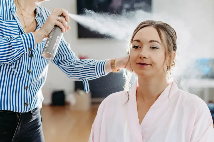 Eine Frau im rosa Morgenmantel lässt sich von einer Person in blau-weiß gestreiftem Hemd die Haare stylen und sprüht dabei gekonnt Haarspray auf. In dem hellen Raum, in dem eine entspannte und konzentrierte Atmosphäre herrscht.