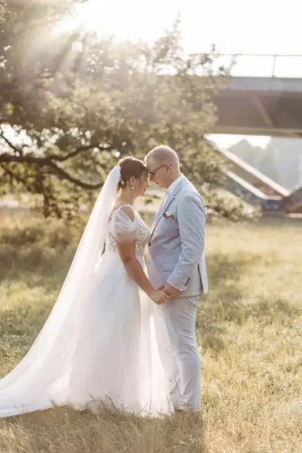 Braut und Bräutigam stehen Hand in Hand auf der Wiese und blicken sich ineinander. Das weiße Kleid und der lange Schleier der Braut flattern leicht, während der hellgraue Anzug des Bräutigams ihre Eleganz unterstreicht. Sonnenlicht fällt durch einen Baum und verleiht diesem Moment, eingefangen von einem Hochzeitsfotografen in Marcolinis Welt, eine romantische Note.