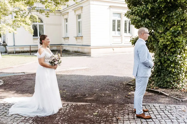 Eine Braut im weißen Kleid mit einem Blumenstrauß in der Hand nähert sich ihrem Bräutigam im hellgrauen Anzug. Sie stehen draußen auf einem Kopfsteinpflasterweg in der Nähe eines prächtigen Gebäudes, umgeben von üppigem Grün. Ein Hochzeitsfotograf in Marcolinis Welt hat das Bild wunderschön festgehalten.