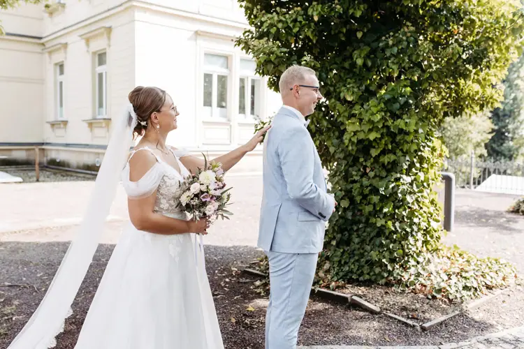 In der bezaubernden Kulisse von Marcolinis Welt berührt eine Braut im weißen Kleid zart die Schulter ihres Bräutigams, der einen hellblauen Anzug trägt. Vor der Kulisse eines üppigen Baumes und eines eleganten Gebäudes wird dieser Moment von einem Hochzeitsfotografen wundervoll festgehalten.