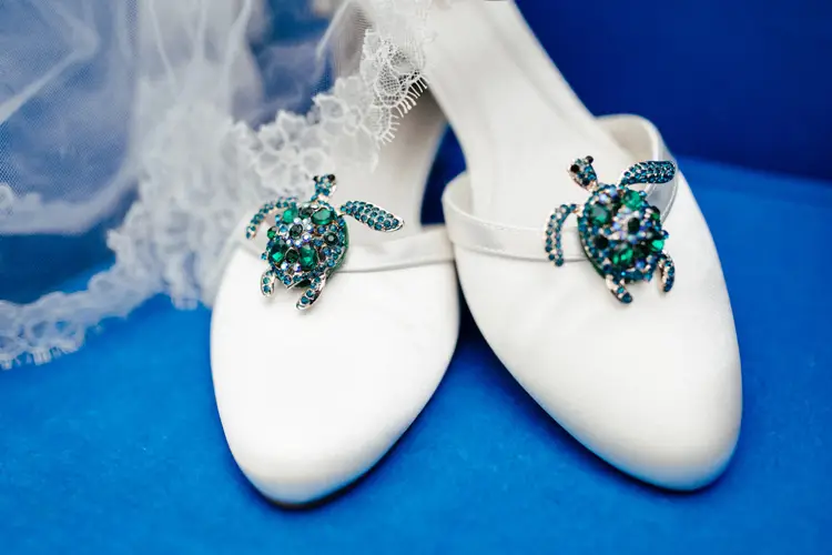 Weiße Brautschuhe, deren dekorative Schildkrötenbroschen mit blauen und grünen Edelsteinen das Licht einfangen, ruhen elegant auf einer leuchtend blauen Oberfläche. Zarter Spitzenstoff blitzt im Hintergrund hervor – eine perfekte Szene für jeden Hochzeitsfotografen in Marcolinis Welt.