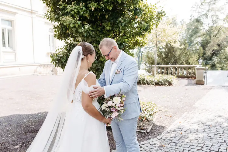 Braut und Bräutigam lächeln sich im Freien freudig an, eingefangen von einem Hochzeitsfotografen in Marcolinis Welt. Die Braut, strahlend in einem schulterfreien Kleid und Schleier, hält ihren Brautstrauß, während der Bräutigam sie in einem hellblauen Anzug begleitet. Sie stehen auf einem Kopfsteinpflasterweg, umgeben von üppigem Grün.