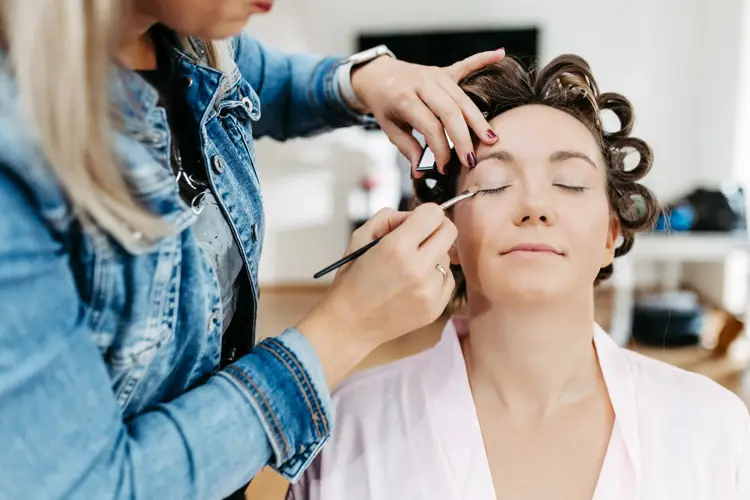 In einem Salon im Innenbereich, entspannt sich eine gelassene Person mit Lockenwicklern, während ihr sorgfältig Augen-Make-up aufgetragen wird und ein Moment festgehalten wird.