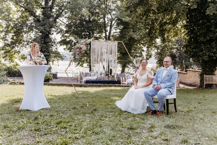 Braut und Bräutigam sitzen während ihrer Hochzeitszeremonie auf einer Bank im Freien, aufgenommen von einem Hochzeitsfotografen in Marcolinis Welt. Die Braut trägt ein weißes Kleid, der Bräutigam einen hellblauen Anzug. In der Nähe steht eine Frau auf einem Podium, eingerahmt von eleganten Bäumen und schimmerndem Wasser.