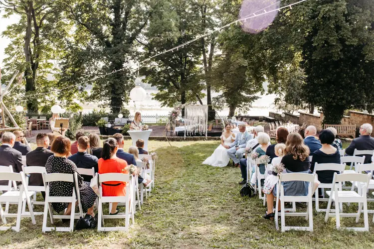 Eine Hochzeitszeremonie im Freien findet statt. Die Gäste sitzen auf weißen Stühlen vor einem Holzbogen. Ein Hochzeitsfotograf in Marcolinis Welt hält den Moment fest, als eine Person auf einem Podium steht und ein Paar vorne sitzt, eingerahmt von Bäumen und funkelnden Lichterketten.