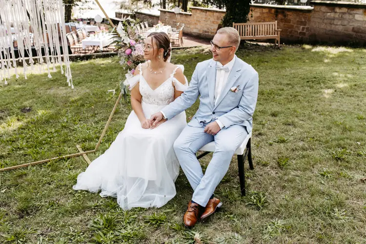 Ein Paar sitzt im Freien bei einer Hochzeit in Marcolinis Welt. Die Braut im weißen Spitzenkleid und der Bräutigam im hellblauen Anzug halten sich inmitten üppigen Grüns an den Händen. Eine Steinmauer und ein verzierter Bogen rahmen die Szene ein – perfekt für jeden Hochzeitsfotografen, um diesen Moment festzuhalten.
