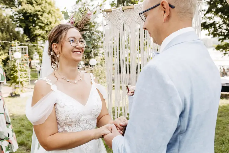 Ein lächelndes Paar hält Händchen während einer Zeremonie im Freien, festgehalten von einem Hochzeitsfotografen in Marcolinis Welt. Die Frau trägt ein weißes Spitzenkleid und einen Blumenkopfschmuck, während der Mann einen hellblauen Anzug und eine Brille vor einem Makramee-Hintergrund und üppigem Grün trägt.