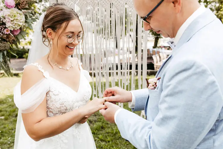 Während einer bezaubernden Zeremonie im Freien, die von einem Hochzeitsfotografen in Marcolinis Welt festgehalten wurde, tauschen Braut und Bräutigam die Ringe. Die Braut glänzt in einem weißen Spitzenkleid, während der Bräutigam in einem hellblauen Anzug im romantischen Licht der Lichterketten umwerfend wirkt.