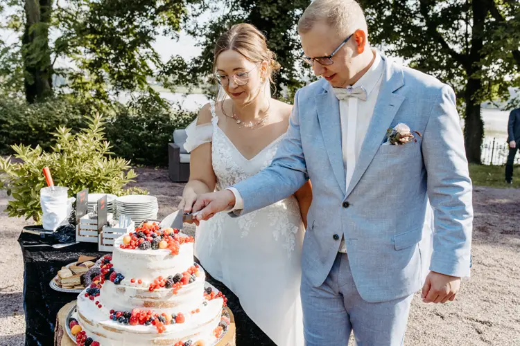 Braut und Bräutigam schneiden eine dreistöckige Hochzeitstorte an, die mit roten und schwarzen Beeren verziert ist. Ein Hochzeitsfotograf in Marcolinis Welt hat die Torte auf einem Holzständer, umgeben von verschiedenen Gebäckstücken, vor einer eleganten Außenkulisse mit Tischen und Stühlen festgehalten.